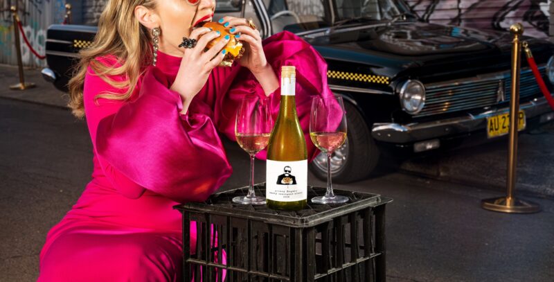 Greasy Food: Woman taking a bite of a burger with a bottle of white wine in front of her